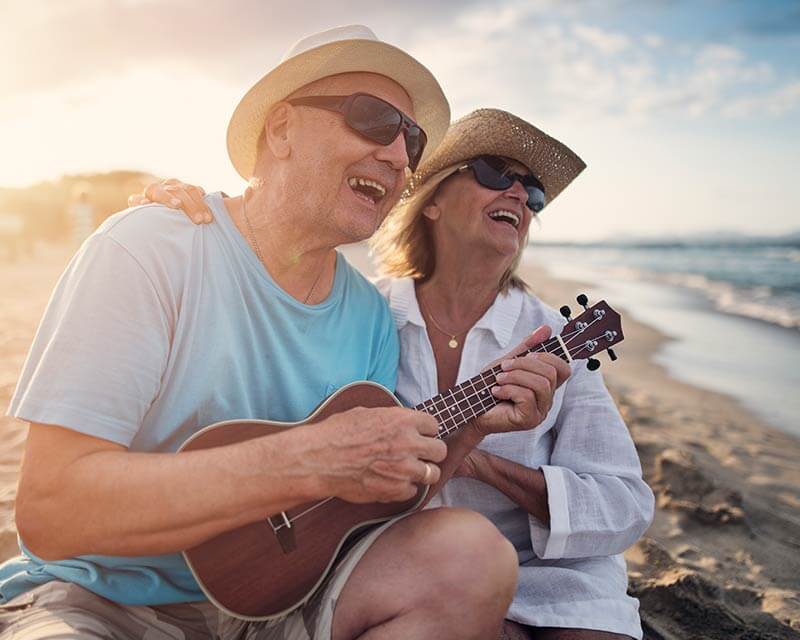 A smiling senior man plays the ukelele with his wife on a beach in Orange County.