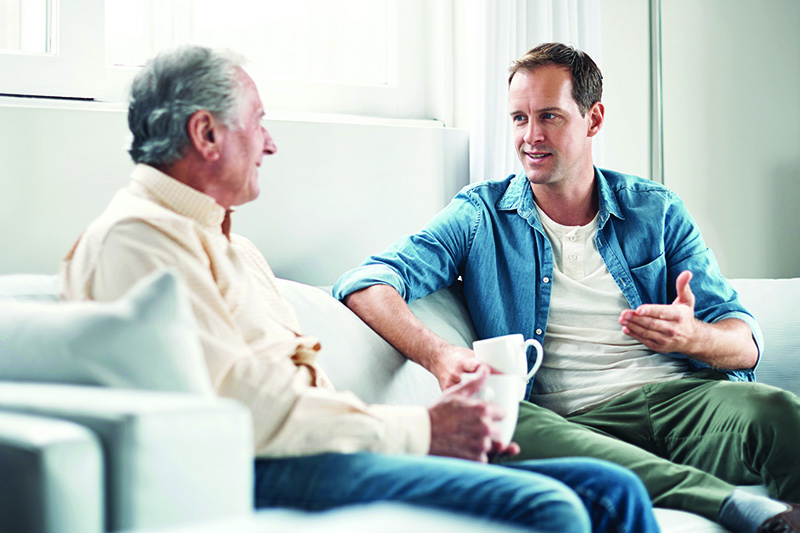 An adult son sits on the couch next to his elderly father drinking coffee and having a chat.