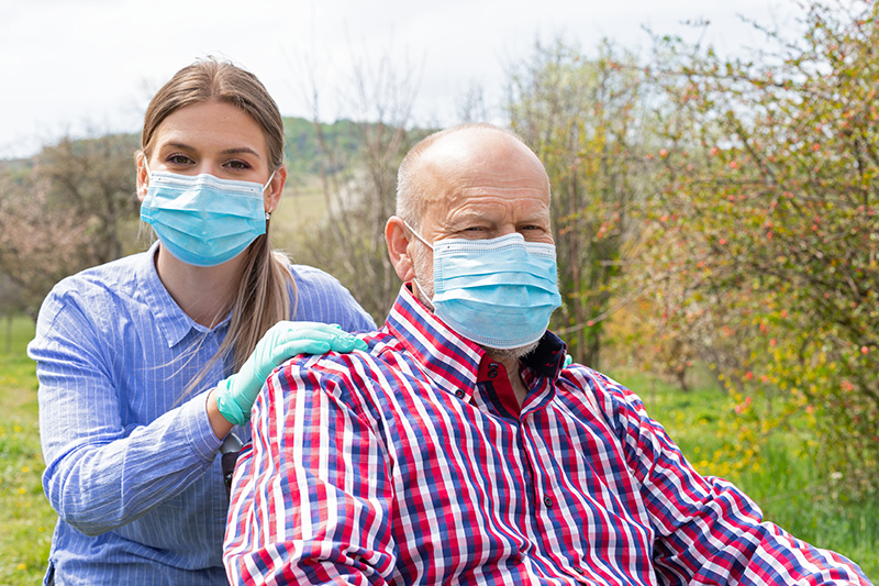 Elderly disabled man with mask sitting in wheelchair, assisted by young female caregiver outdoors