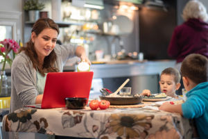 caregiver sitting at the dining table and working on laptop, her two sons are having lunch, grandmother is working in the kitchen