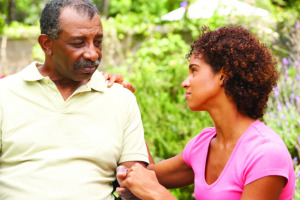 An adult daughter kneels next to her elderly father and holds his hand.