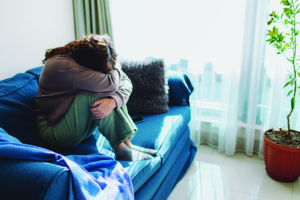 A woman sits on a blue couch with her knees up and her head bowed and covered by her arms.