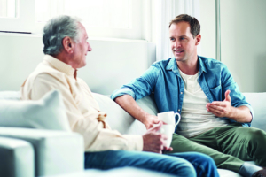 An adult son sits on the couch next to his elderly father drinking coffee and having a chat.