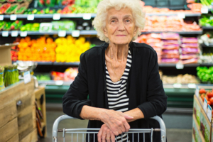 An older woman pushes a grocery cart in the produce section of the store.