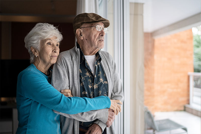 A woman looks out the window while embracing her elderly husband as she grapples with the stress of caring for a spouse.