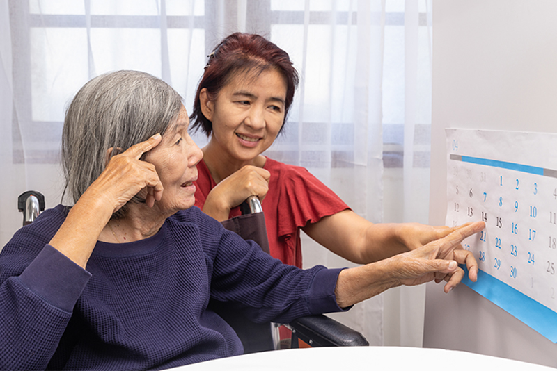 A woman works at managing reality in dementia support with her aging mother by pointing out the date on the calendar.