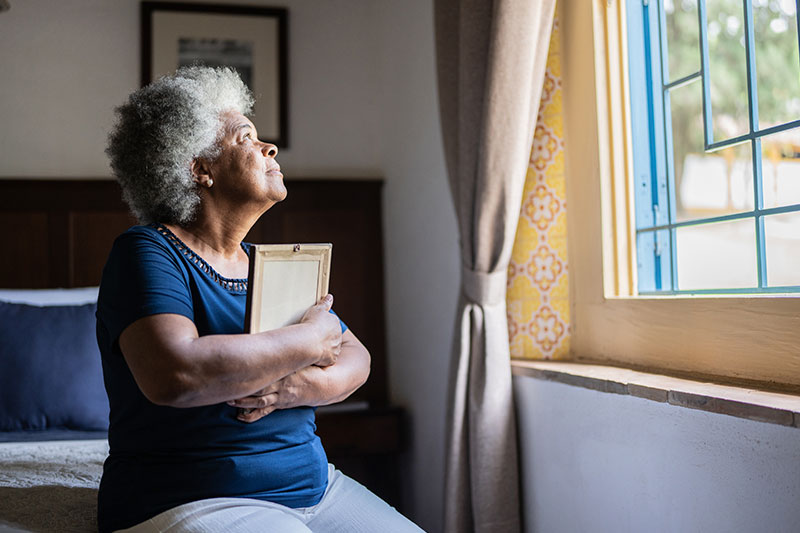 An elderly woman holds a framed picture while staring out the window. Supporting seniors through loss can be an emotional process.