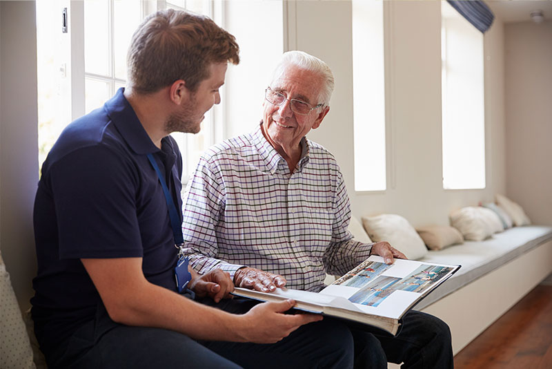 An older man and his dementia care provider sit side by side and look at a photo album together.