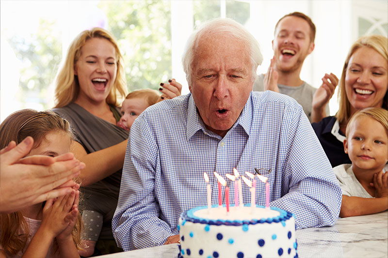A senior man blows out candles on a birthday cake as his family cheers him on. They know the importance of coming up with birthday party ideas for seniors to ensure their loved one feels special.