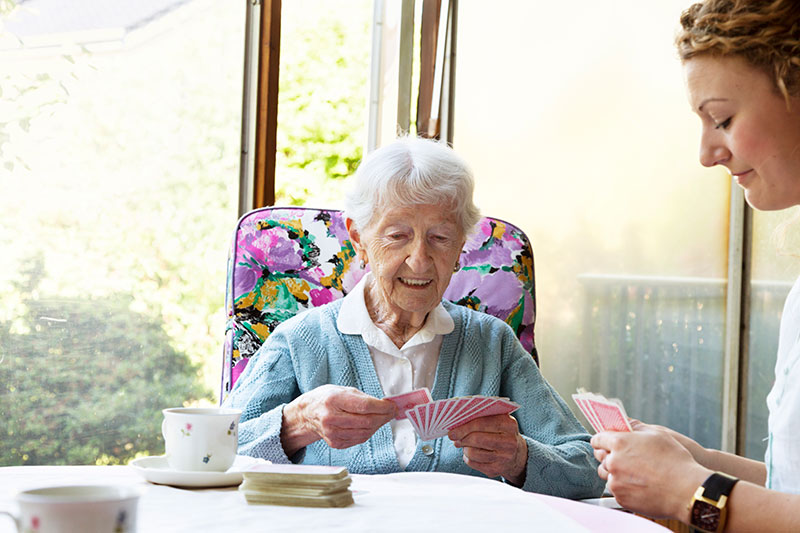 An older woman is smiling and playing cards with her companion care provider in Huntington Beach.