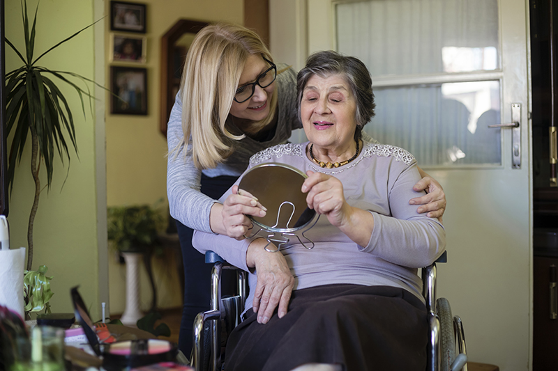 An older woman in Huntington Beach sits in a wheelchair holding a small mirror, smiling as she looks at her reflection with her personal care provider behind her.